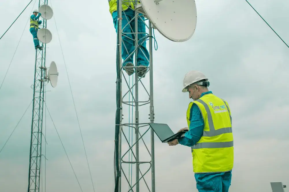 Technicians performing telecommunications tower maintenance, with one using a laptop to run field service management software from Appenate.