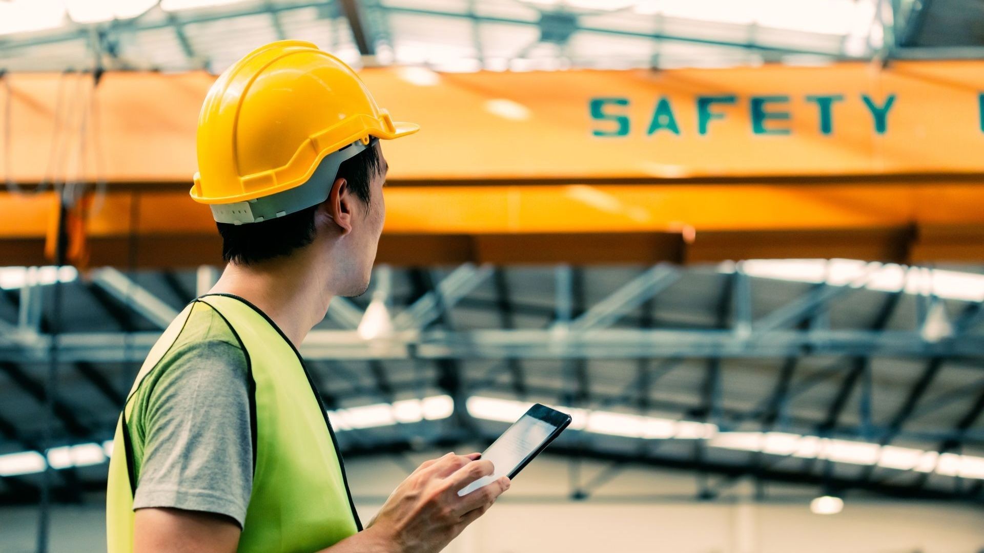 A man wearing a hard hat and safety vest is looking at a tablet.
