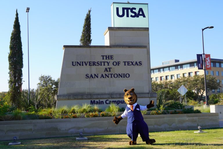 Alan CyBear posing in front of the UT San Antonio main campus entrance sign